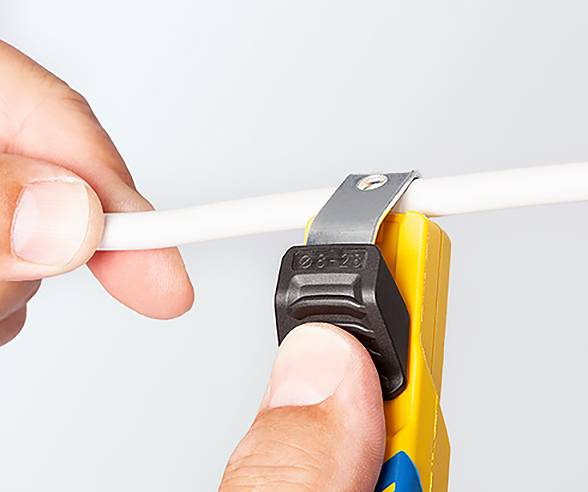 Eine Person verwendet ein Utility-Messer mit einem gelben Griff, um ein weißes Kabel zu durchtrennen. Das Messer verfügt über eine schwarze, verstellbare Klingenschutzabdeckung.