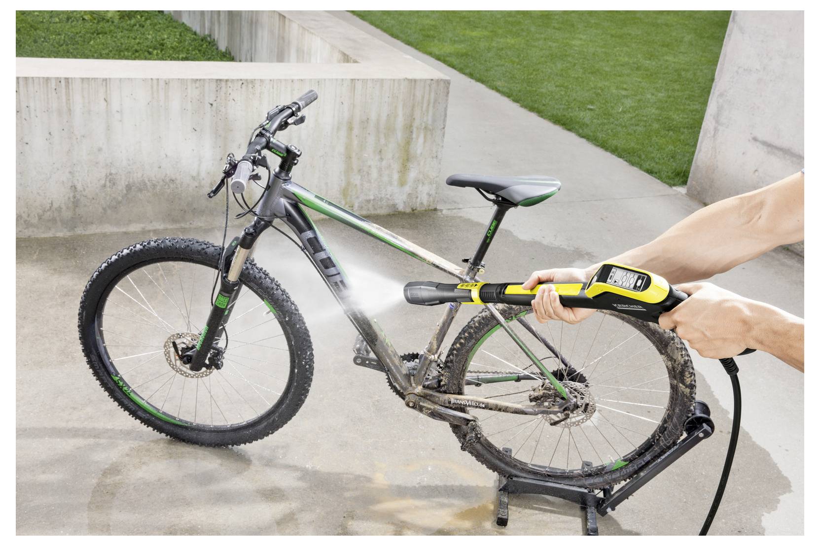 Eine Person benutzt einen Hochdruckwasserstrahl, um ein schlammverschmutztes Fahrrad auf einer Betonoberfläche zu reinigen, während im Hintergrund ein Grasfläche zu sehen ist.
