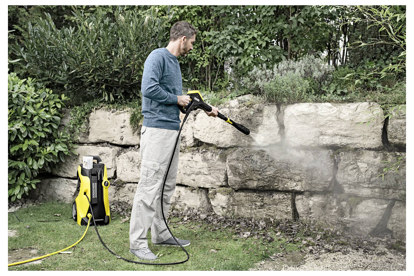 Eine Person verwendet einen Hochdruckreiniger, um eine Steinmauer in einem Garten zu reinigen, umgeben von Grünpflanzen und Büschen.