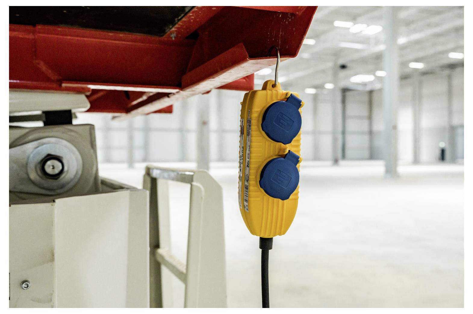 Gelber industrieller Pendelschalter mit blauen Tasten, der von roter Maschine in einer hellen Fabrik oder Werkstatt hängt.