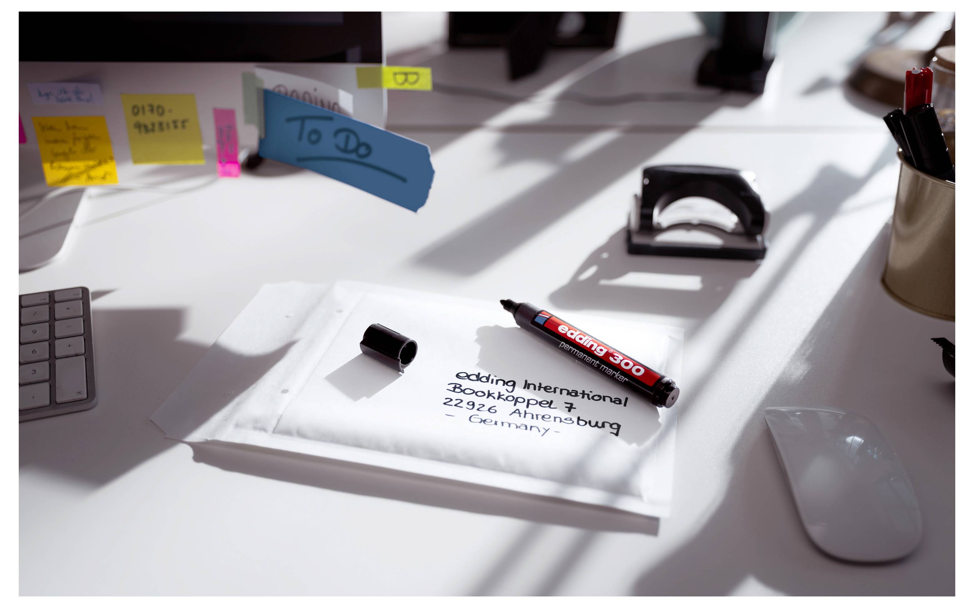 Schreibtisch mit einem gefalteten weißen Paket mit der Aufschrift „booking international" und einem roten Marker darauf, umgeben von Büromaterialien und Haftnotizen.