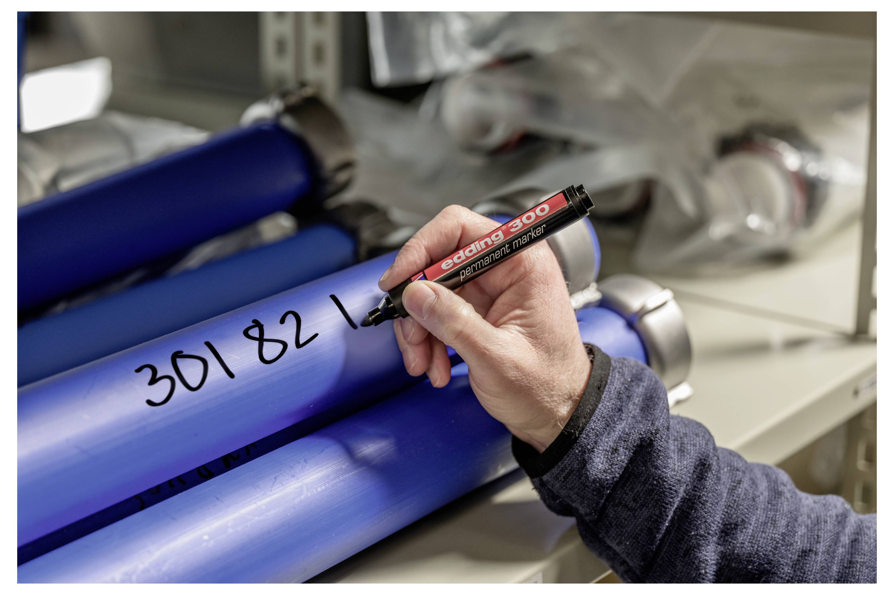 Handschriftlich eine Identifikationsnummer mit einem Marker auf ein blaues Industrierohr in einer Werkstatt- oder Fabrikumgebung schreiben.