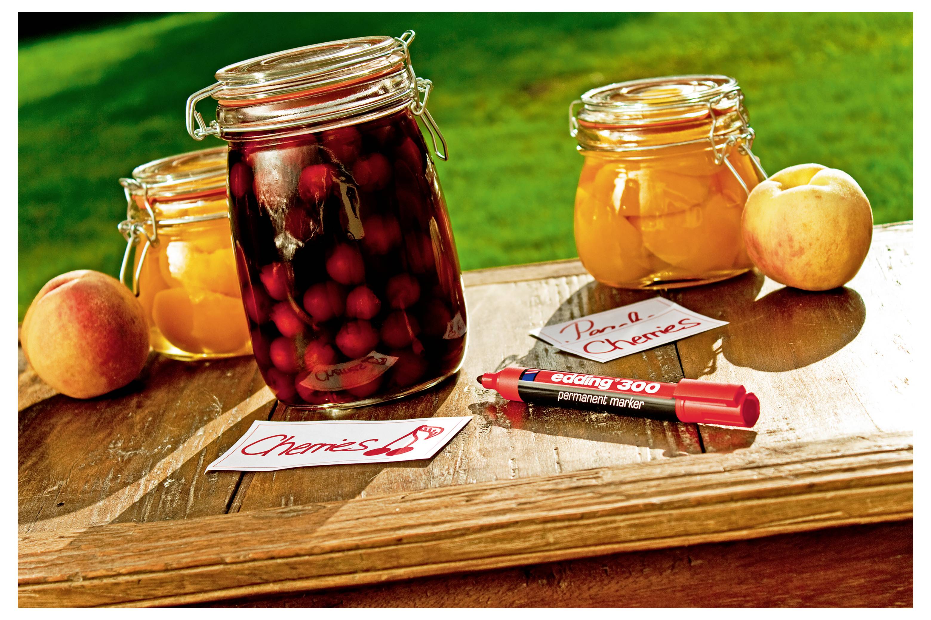 Gläser mit eingelegtem Obst auf einem Holztisch, darunter ein großes Glas mit Kirschen oder Pflaumen und kleinere Gläser mit Pfirsichkonserven sowie frische Pfirsiche in der Nähe.