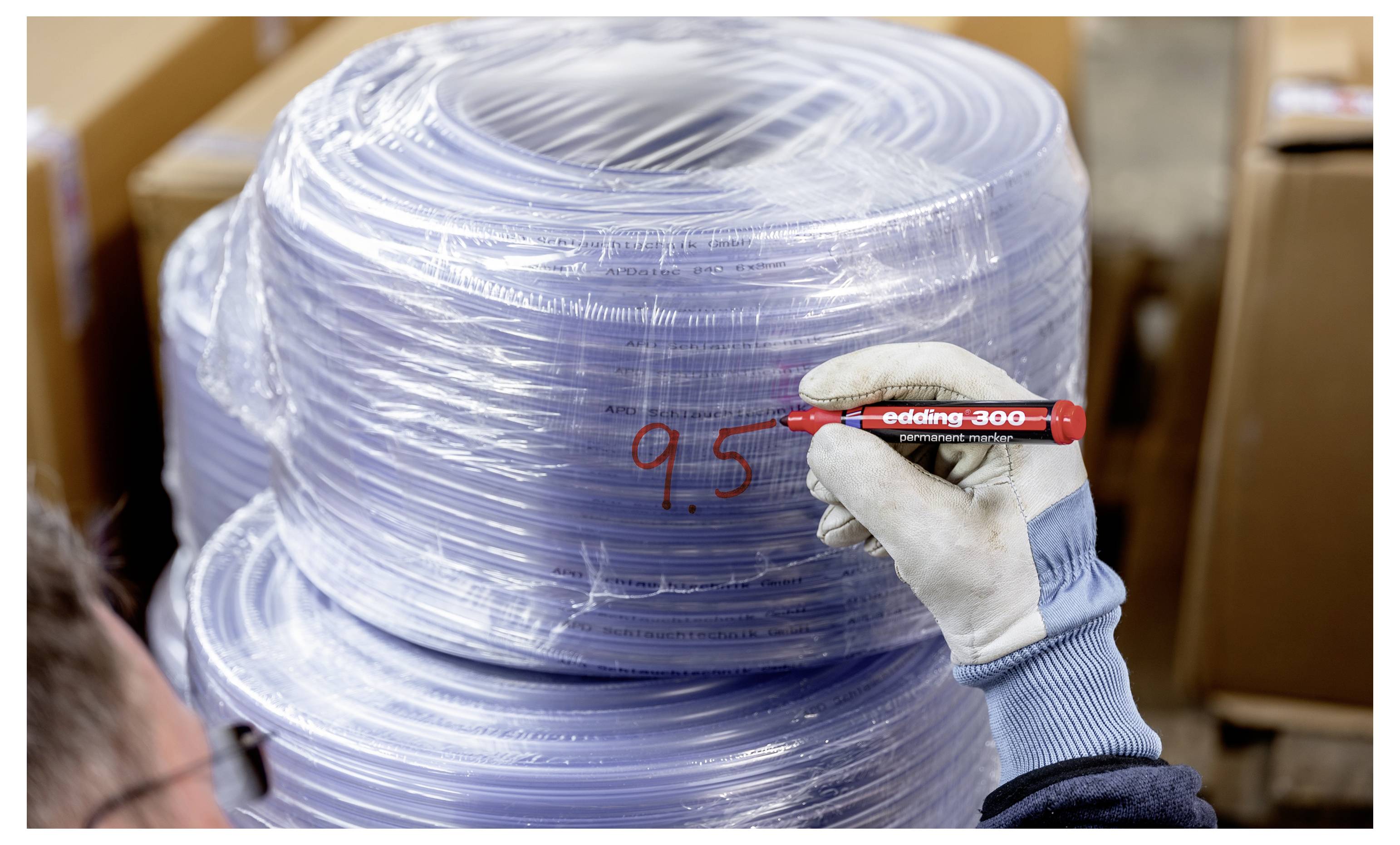 Handschuhgetragener Arbeiter schreibt mit einem roten Marker „9,5" auf eine große aufgerollte Rolle aus transparenter Kunststofffolie.