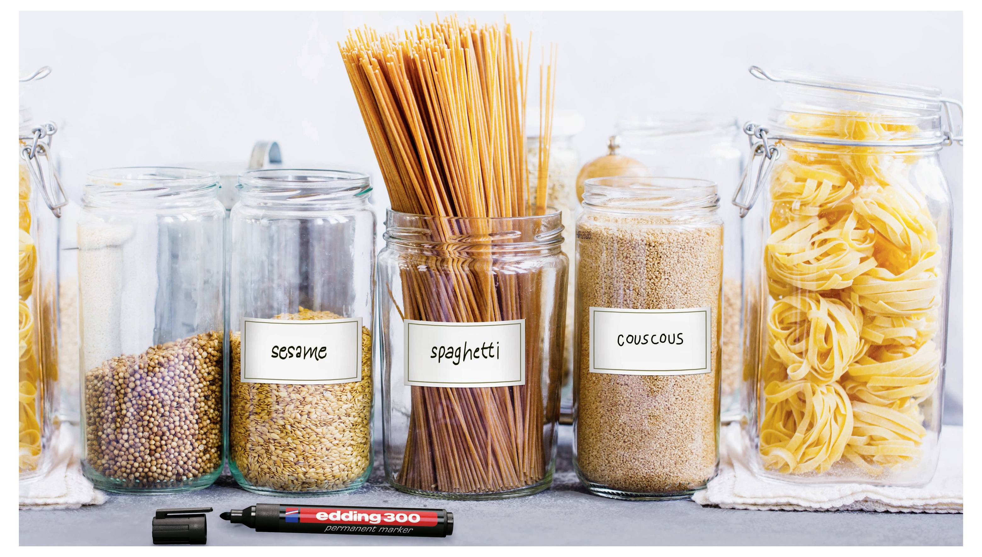 Fünf Glaskäse mit trockenen Lebensmitteln auf einem Regal, etikettiert mit Sesam, Spaghetti, Couscous und Pasta, mit einem entriegelten Marker davor.