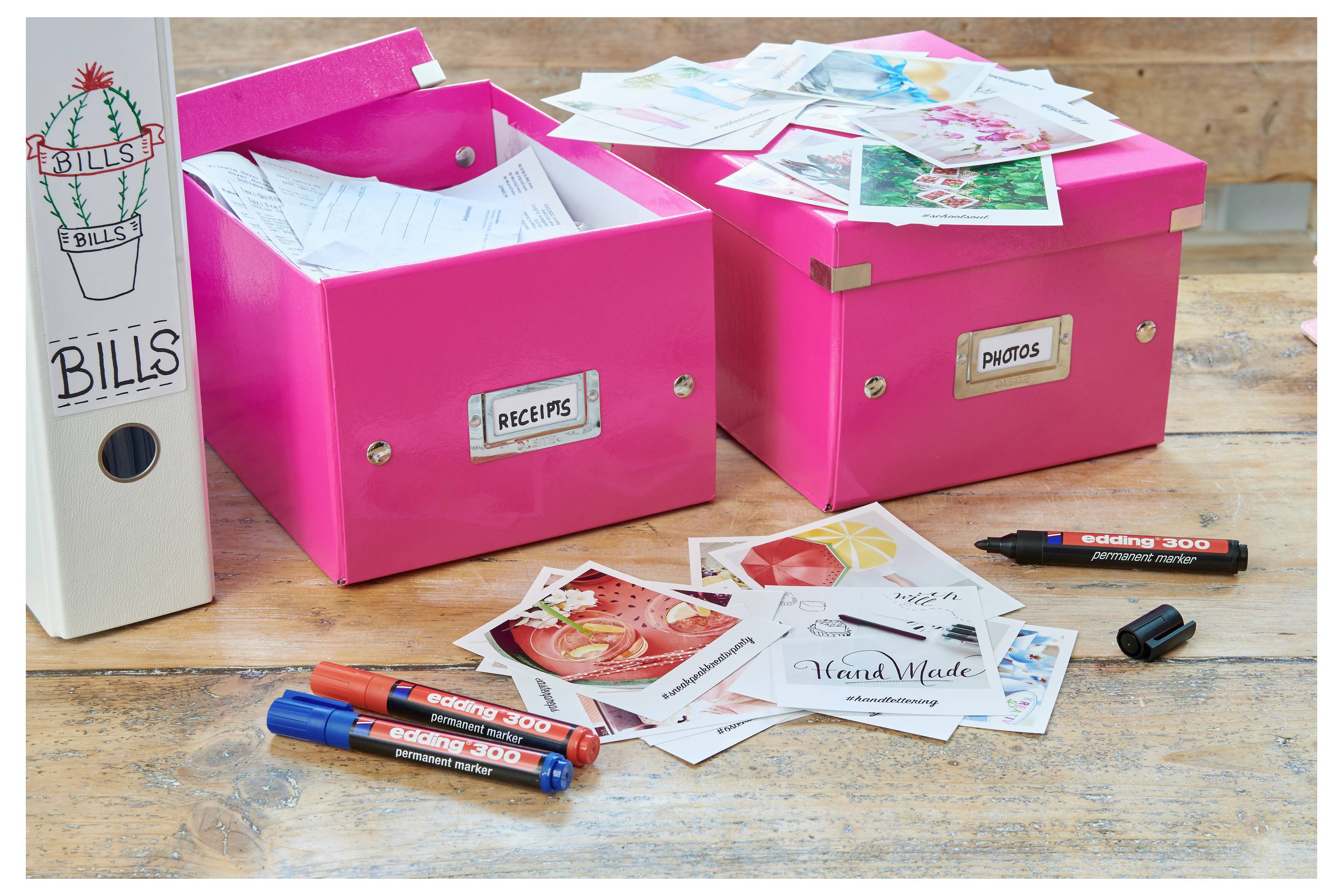 Zwei rosa Aufbewahrungsboxen mit den Beschriftungen „QUITTUNGEN" und „FOTOS" enthalten Papiere und Schnappschüsse, neben einer Datei mit der Beschriftung „RECHNUNGEN" und verstreuten Markern auf einem Tisch.