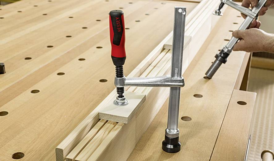 Eine Schraubzwinge klemmt ein Holzstück auf einer Werkbank. Eine Hand hält ein weiteres Werkzeug, möglicherweise für Feinjustierung.