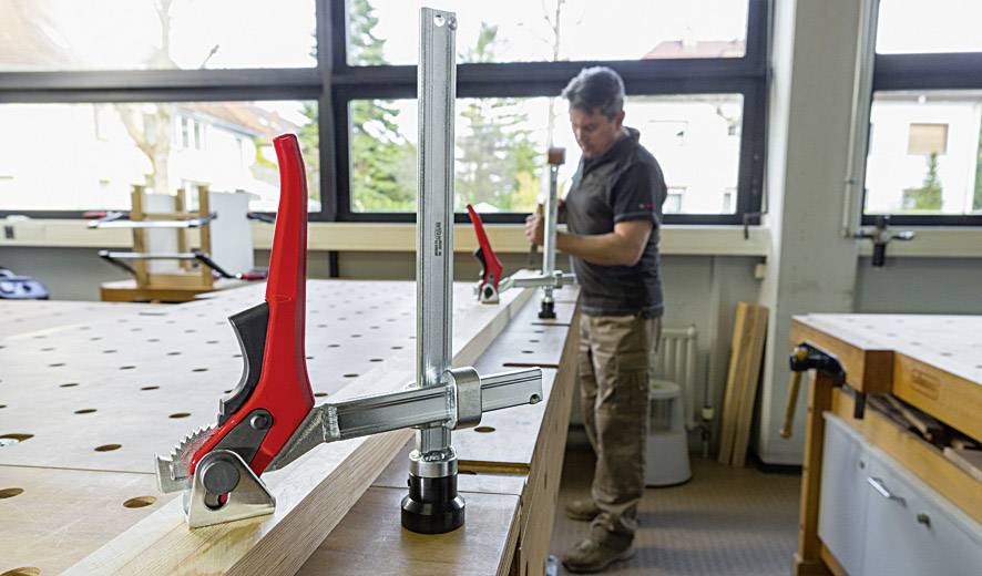 Ein Mann arbeitet in einer Werkstatt, spannt Holzplatten mit roten Klemmen auf einem Tisch fest. Im Hintergrund sind Fenster zu sehen.