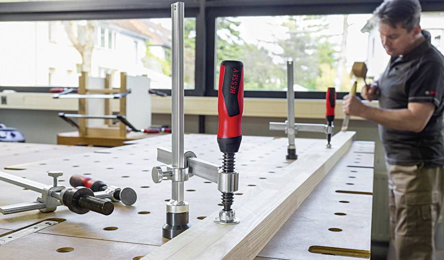 Ein Mann arbeitet in einer Werkstatt, klemmt Holz auf einem Tisch mit mehreren Spannvorrichtungen fest. Helle, gut beleuchtete Umgebung.