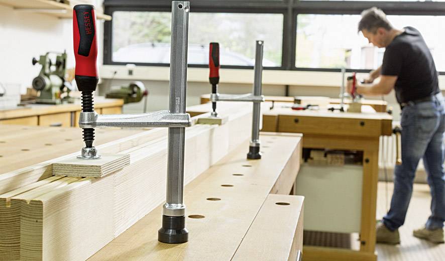 Ein Mann arbeitet in einer Werkstatt an einem Holztisch mit Schraubzwingen. Im Hintergrund sind Werkzeuge und Maschinen zu sehen.