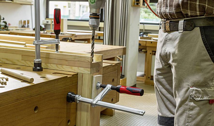 Eine person bohrt mit einer Bohrmaschine in ein Holzstück, das an einem Tisch in einer Werkstatt mit Klammern befestigt ist.