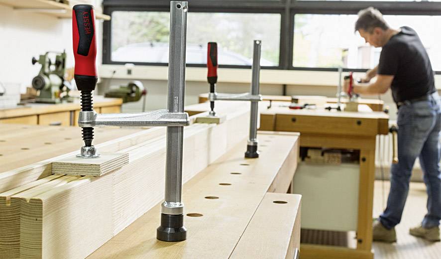 Ein Handwerker arbeitet in einer Werkstatt an einem Holztisch, auf dem mehrere Schraubzwingen befestigt sind. Im Hintergrund sind Maschinen zu sehen.