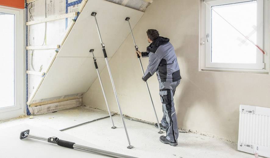 Ein Mann installiert Dämmmaterial an einer schrägen Zimmerdecke mit Teleskopstangen, in einem Raum mit Fenster und Heizkörper.