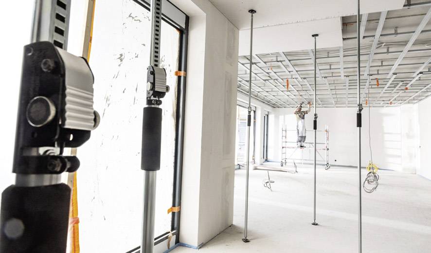 Eine Baustelle mit verstellbaren Stützen, die eine abgehängte Decke tragen. Ein Arbeiter steht im Hintergrund auf einem Gerüst.