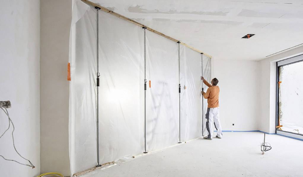 Eine Person zieht eine Plastikfolie an der Wand in einem leeren Raum straff. Möglicherweise eine Renovierungsszene.