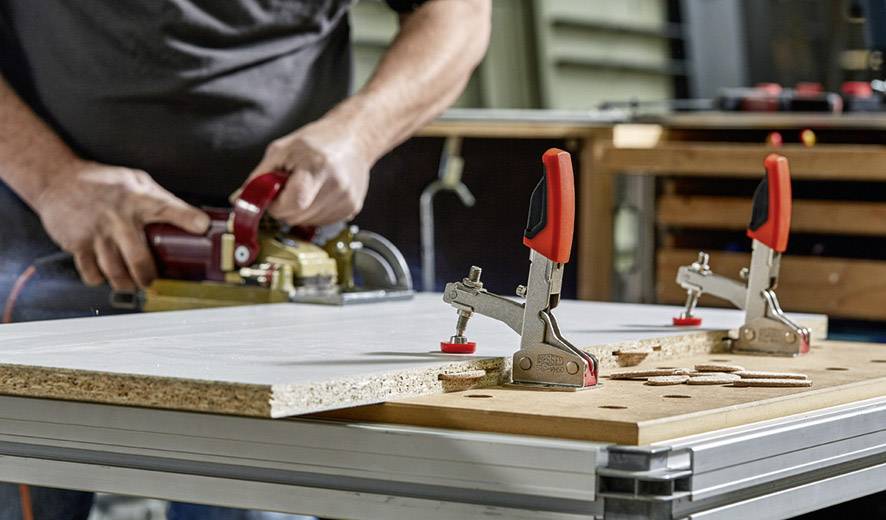 Eine Person schleift eine Holzplatte, die mit zwei Schraubzwingen auf einem Arbeitstisch befestigt ist. Im Hintergrund sind Werkzeuge sichtbar.