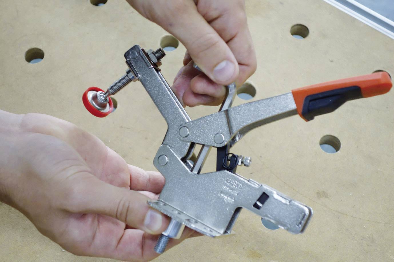 Eine Person hält eine Metallklemme mit roten und schwarzen Griffen über einem Holztisch mit Löchern.