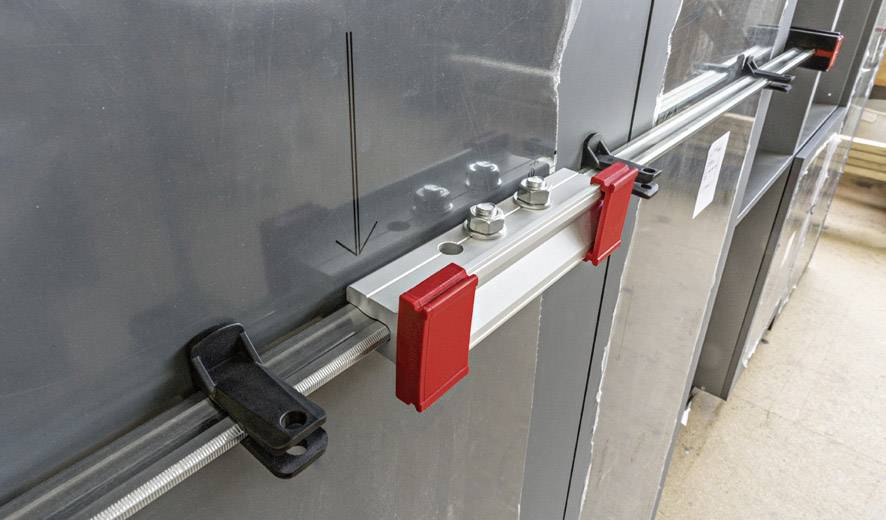 Eine Klemme wird an einem grauen Metallpaneel befestigt. Rote und schwarze Komponenten sichern die Verbindung. Eine schwarze Pfeilmarkierung zeigt die Richtung an.
