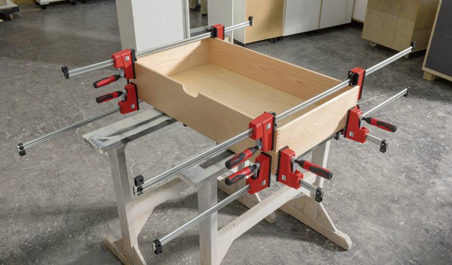 Holzschublade auf Werkbank, mit sechs großen Schraubzwingen fixiert. Schraubzwingen sind rot und halten die Teile beim Trocknen zusammen.