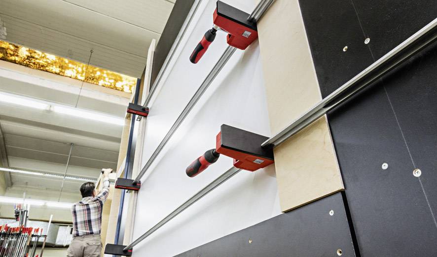 Eine Person befestigt Holzplatten in einer vertikalen Klemmanlage in einer Werkstatt. Die Klemmen sind rot und schwarz.