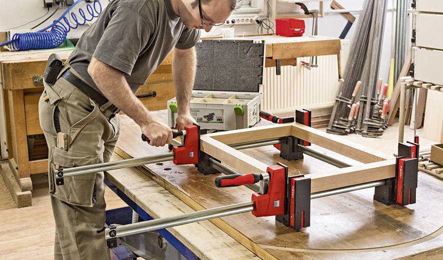 Ein Mann arbeitet in einer Werkstatt und verwendet Zwingen, um Rahmenteile auf einem Tisch zu fixieren. Werkzeuge und Materialien sind um ihn herum.