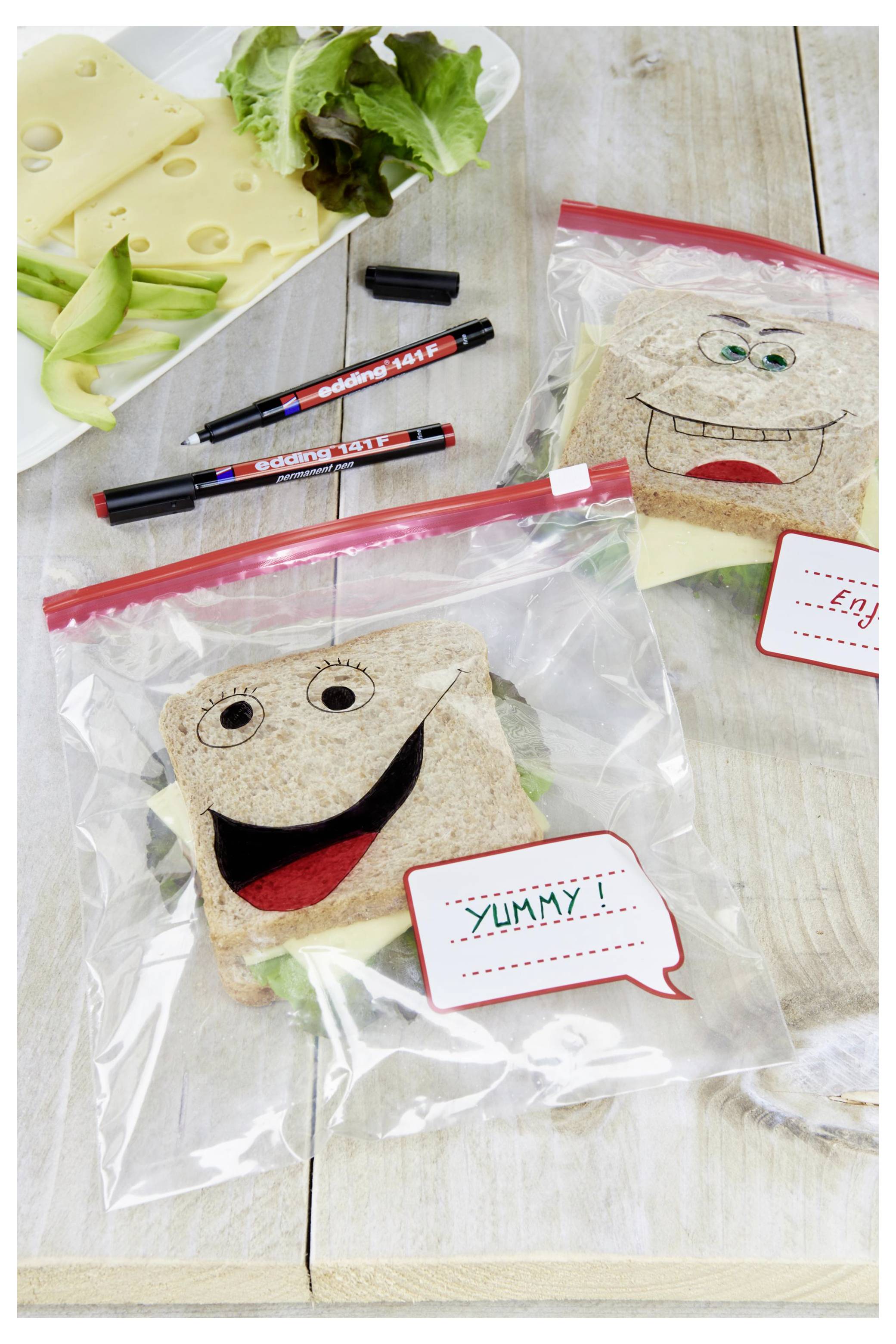 Zwei lächelnde Sandwich-Gesichter in transparenten Reißverschlussbeuteln auf einem Tisch, mit Käse, Salat und Markern in der Nähe zum Verzieren von Gesichtern auf den Sandwiches.