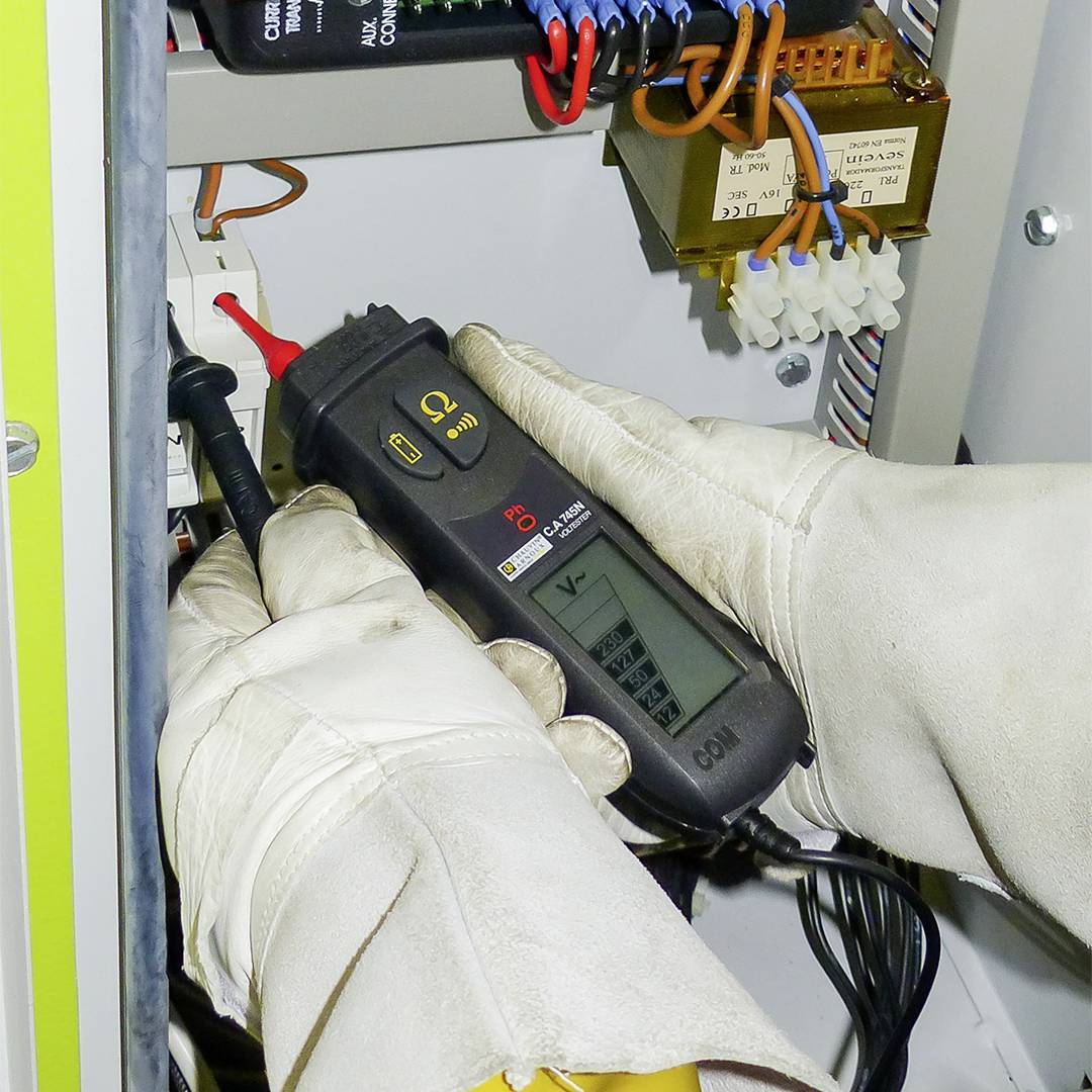 Ein Techniker misst elektrische Spannung in einem Schaltschrank mit einem Spannungsprüfer. Kabel und Bauteile sind sichtbar.