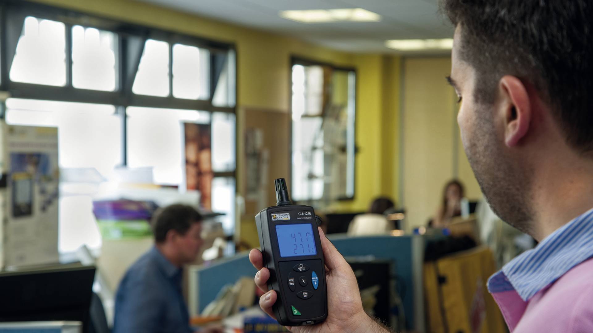 Ein Mann hält ein Luftqualitätsmessgerät in einem Büro, im Hintergrund arbeiten Menschen an Schreibtischen.