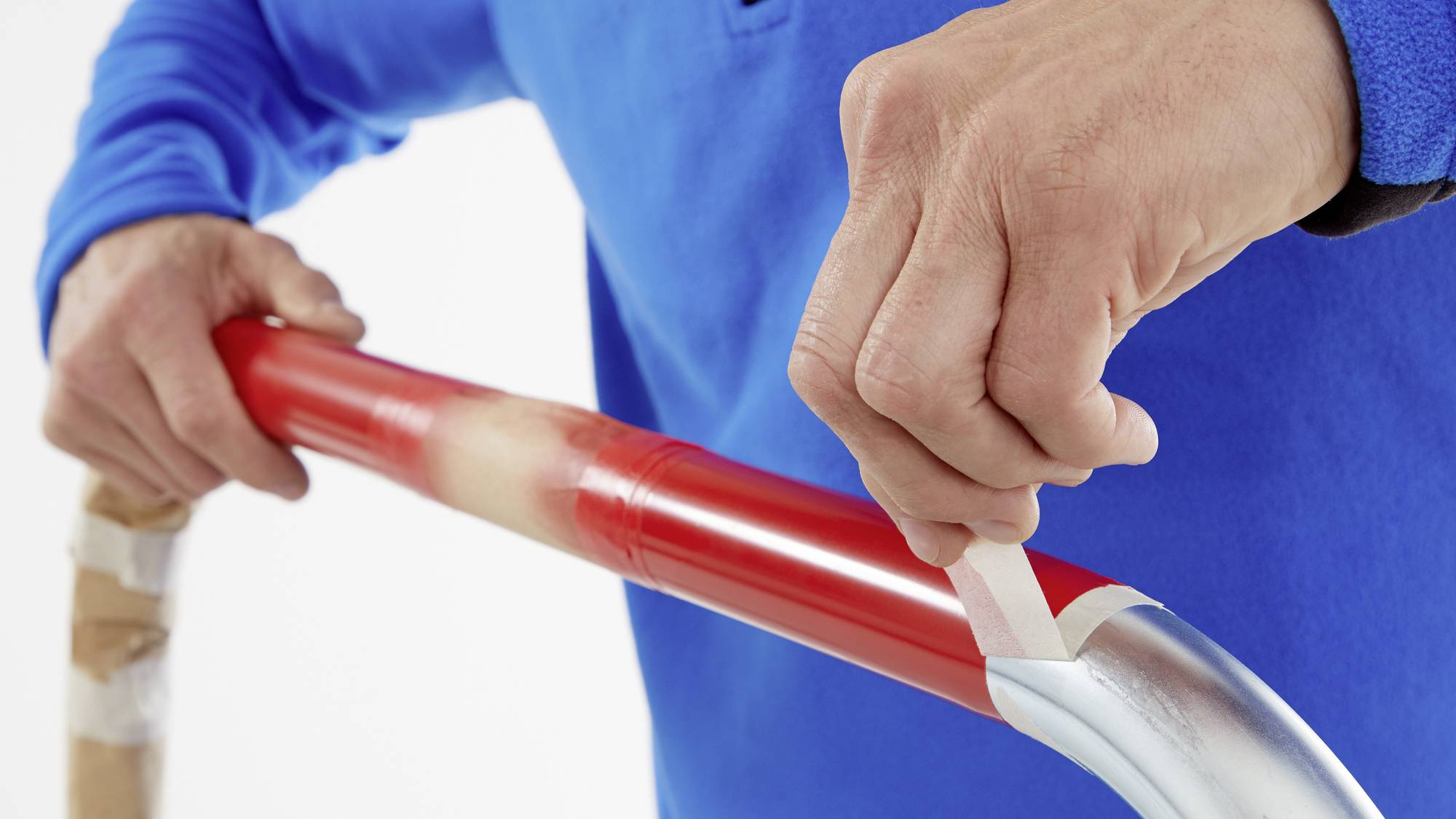 Eine Person in blauem Pullover klebt ein rotes Hockeyschlägerrohr mit weißem Tape ab. Händen fokussiert auf präzise Wicklung.