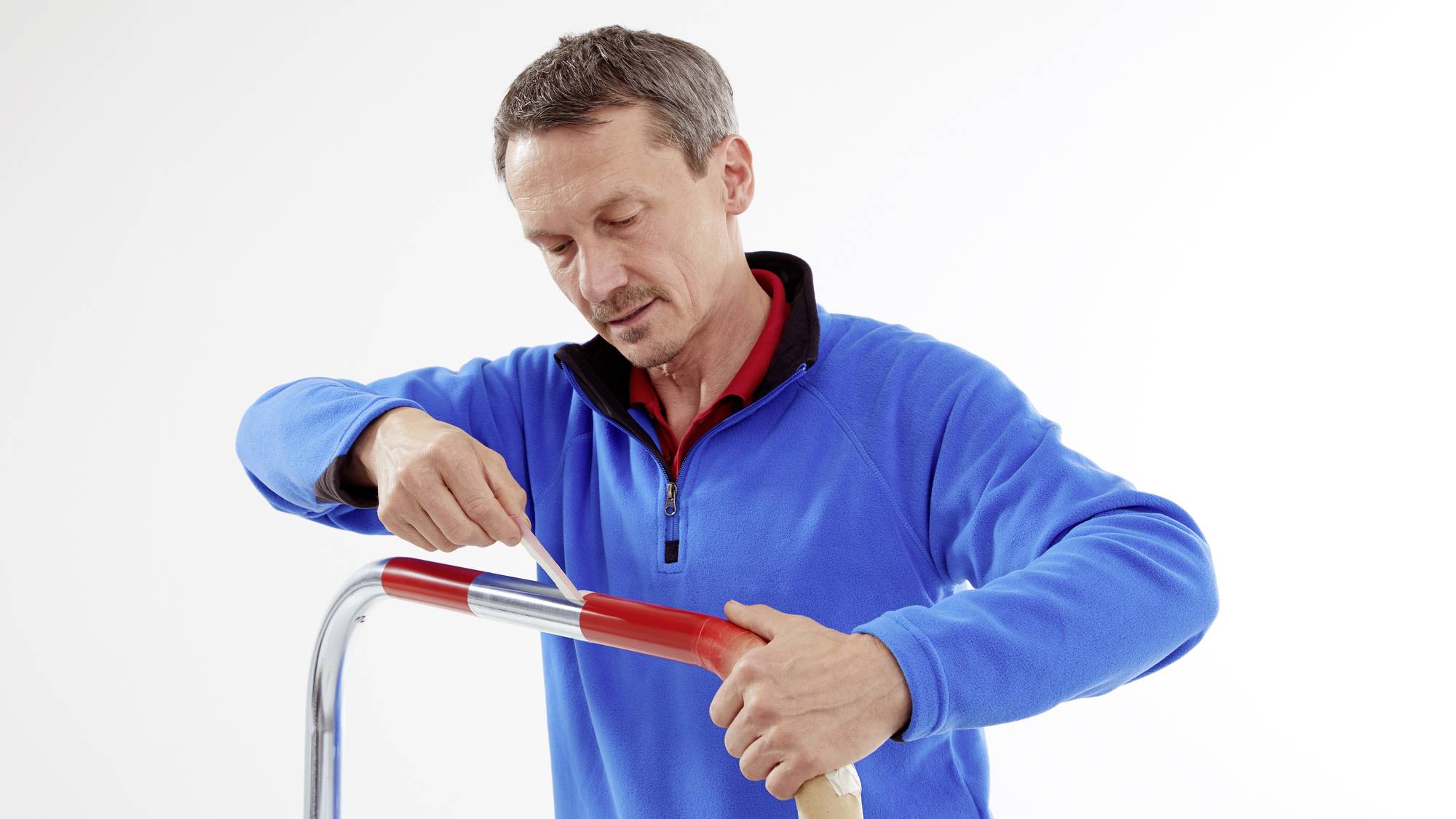 Ein Mann in einem blauen Pullover verwendet ein Teppichmesser, um das Ende eines roten Isolierbands an einer Metallstange abzuschneiden.