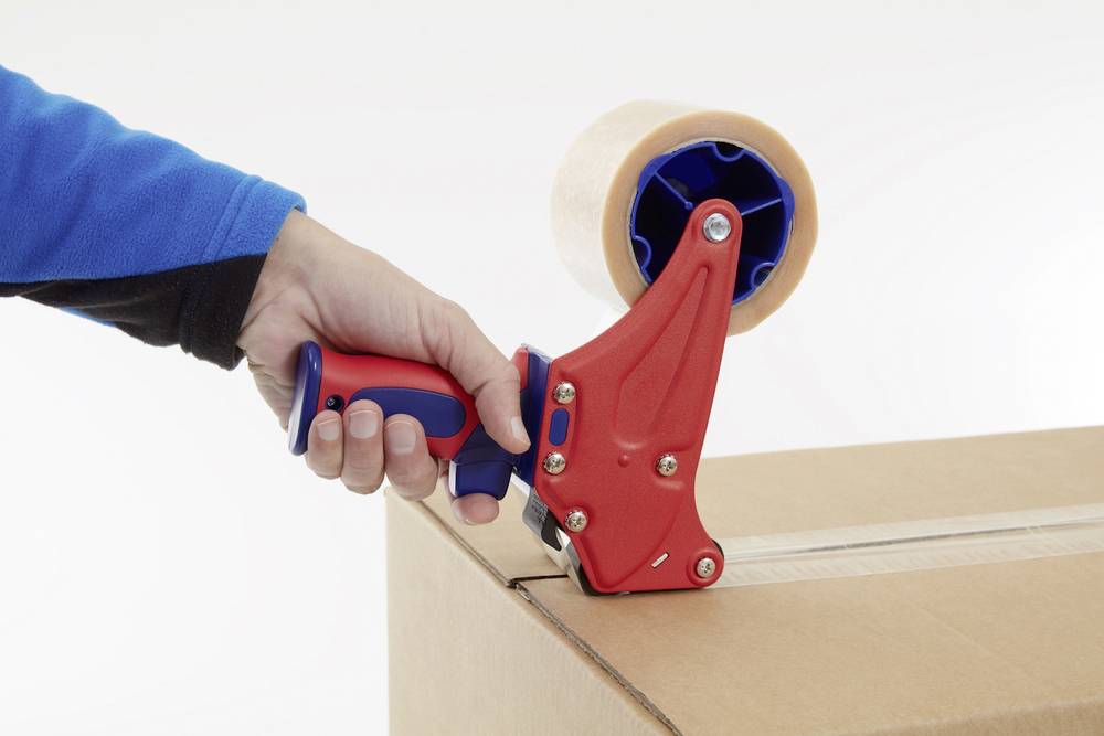 Eine Person verschließt einen Karton mit einem roten Handabroller für Klebeband, der auf der Kartonecke angesetzt wird.
