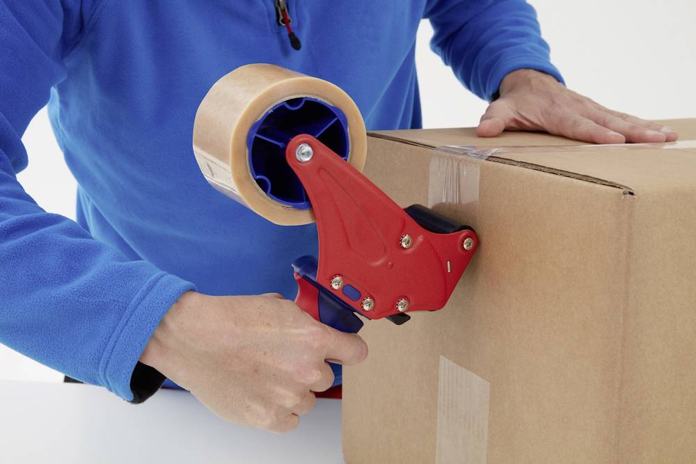 Person verpackt einen Karton mit Klebeband und einem Bandabroller. Hände in blauer Jacke arbeiten präzise.