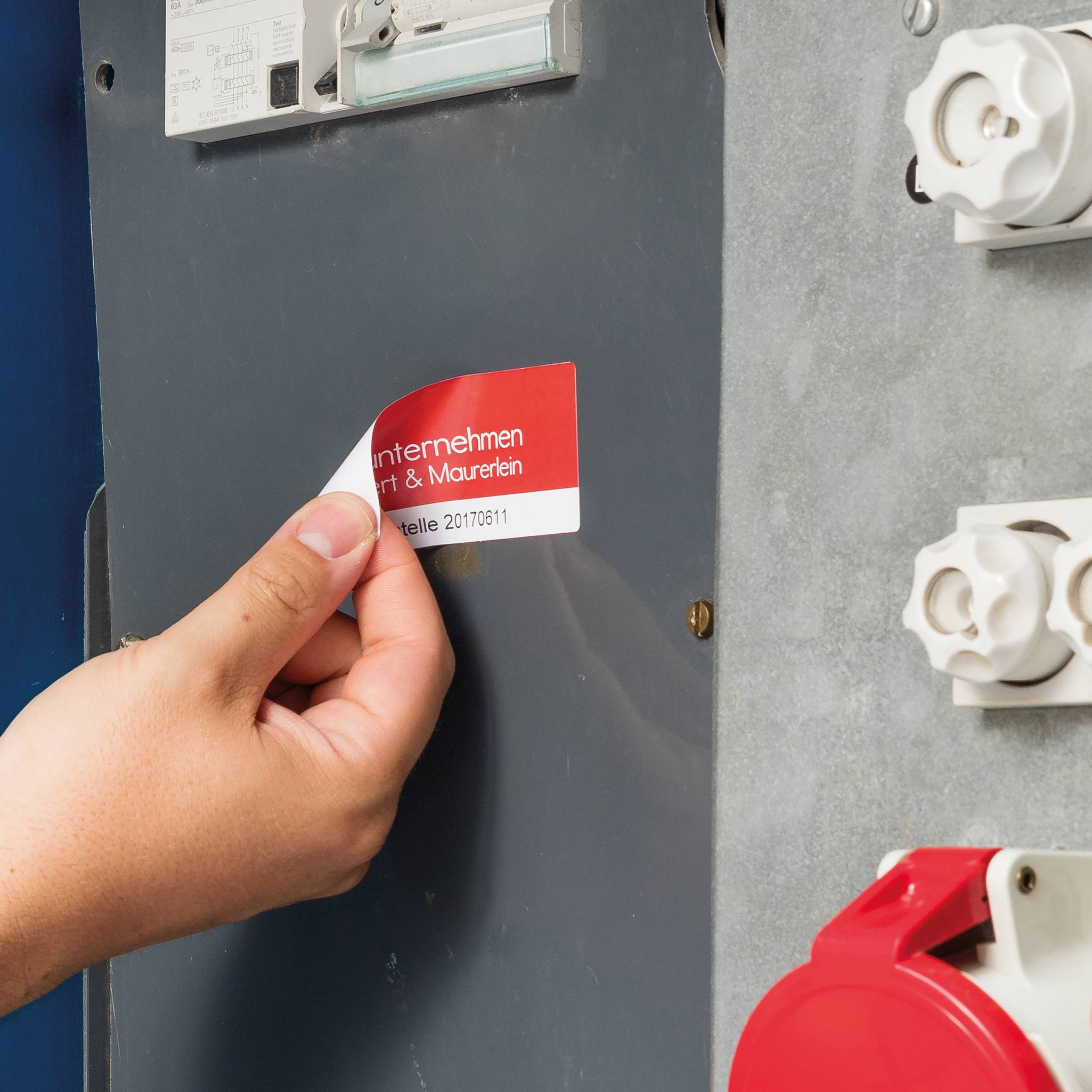 Eine Hand entfernt ein rotes Etikett von einem grauen Schaltschrank. Auf dem Etikett steht 'unternehmen | Maler & Maurer'.