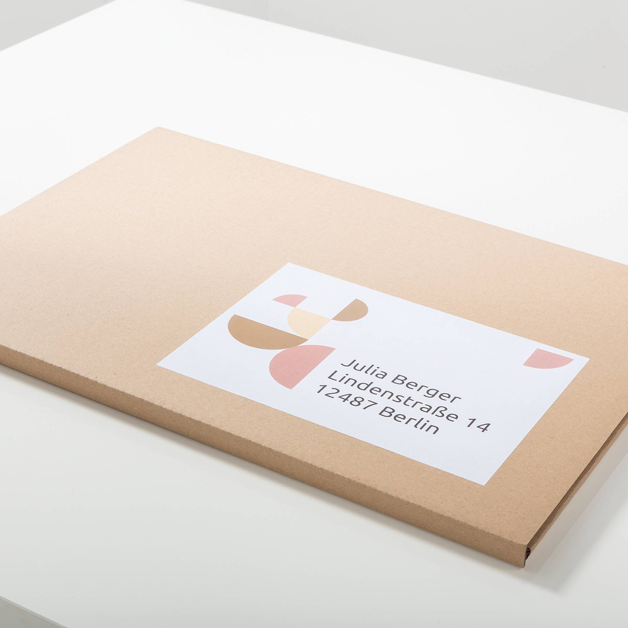 Ein rechteckiger, flacher, brauner Kartonumschlag mit einem Aufkleber, der bunte Formen und die Adresse 'Julia Berger, Lindenstraße 14, 12487 Berlin' zeigt.