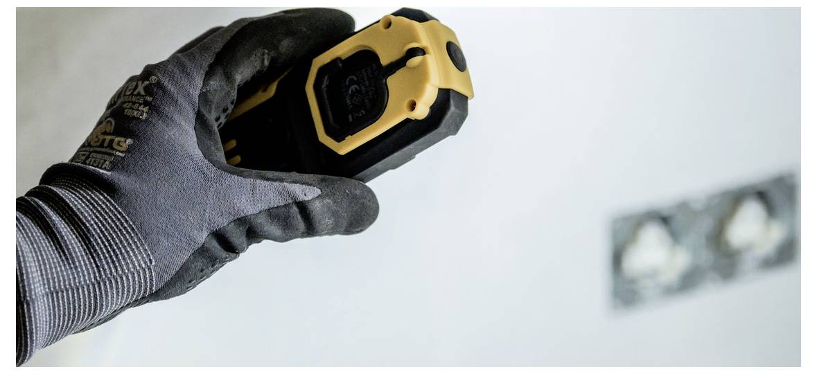 Behandschuhte Hand, die ein gelbes Laser-Entfernungsmessgerät auf eine Wand richtet, was auf Mess- oder Layoutarbeiten während einer Renovierung hindeutet.