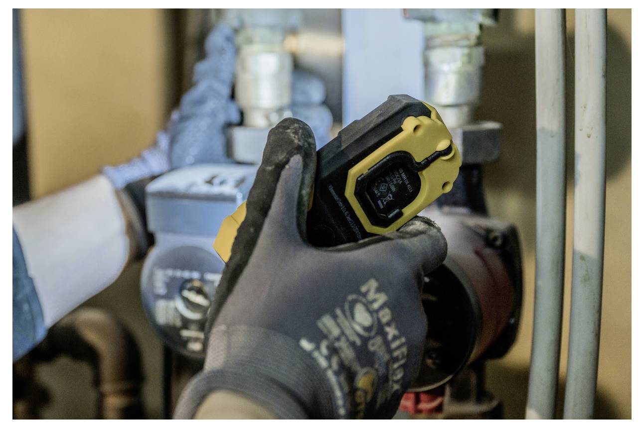 Handschuhgetragener Arbeiter, der ein gelbes Infrarot-Thermometer verwendet, um ein Rohr oder einen Motor in einem industriellen Versorgungsbereich zu überprüfen.