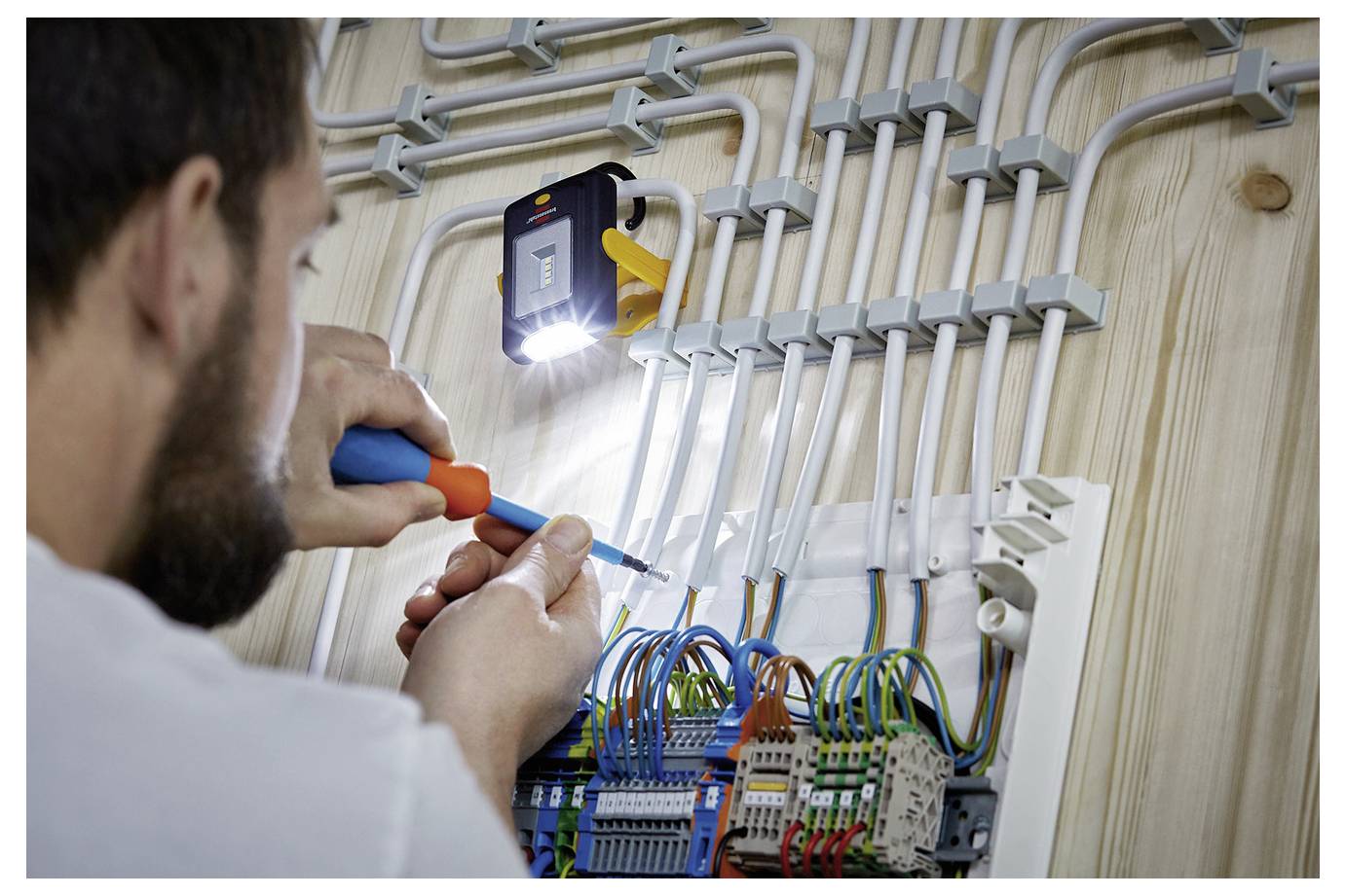 Elektriker verdrahtet ein Elektroschaltfeld, verbindet farbige Drähte in einer wandmontierten Verteilertafel, während er eine Taschenlampe zur Sichtbarkeit benutzt.