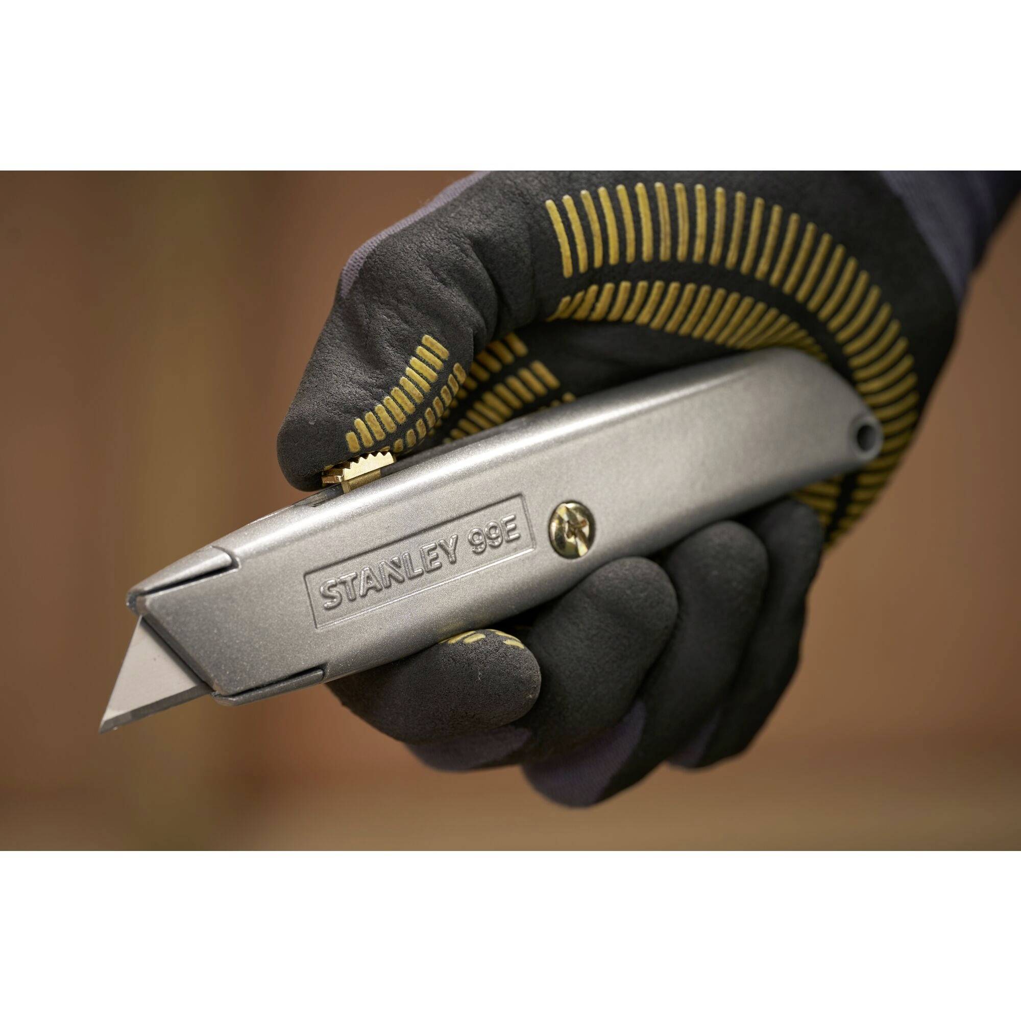 Eine Person hält ein silbernes Stanley 99E Universalmesser mit einer Klinge in einer schwarzen, gelb gemusterten Arbeitshandschuh.