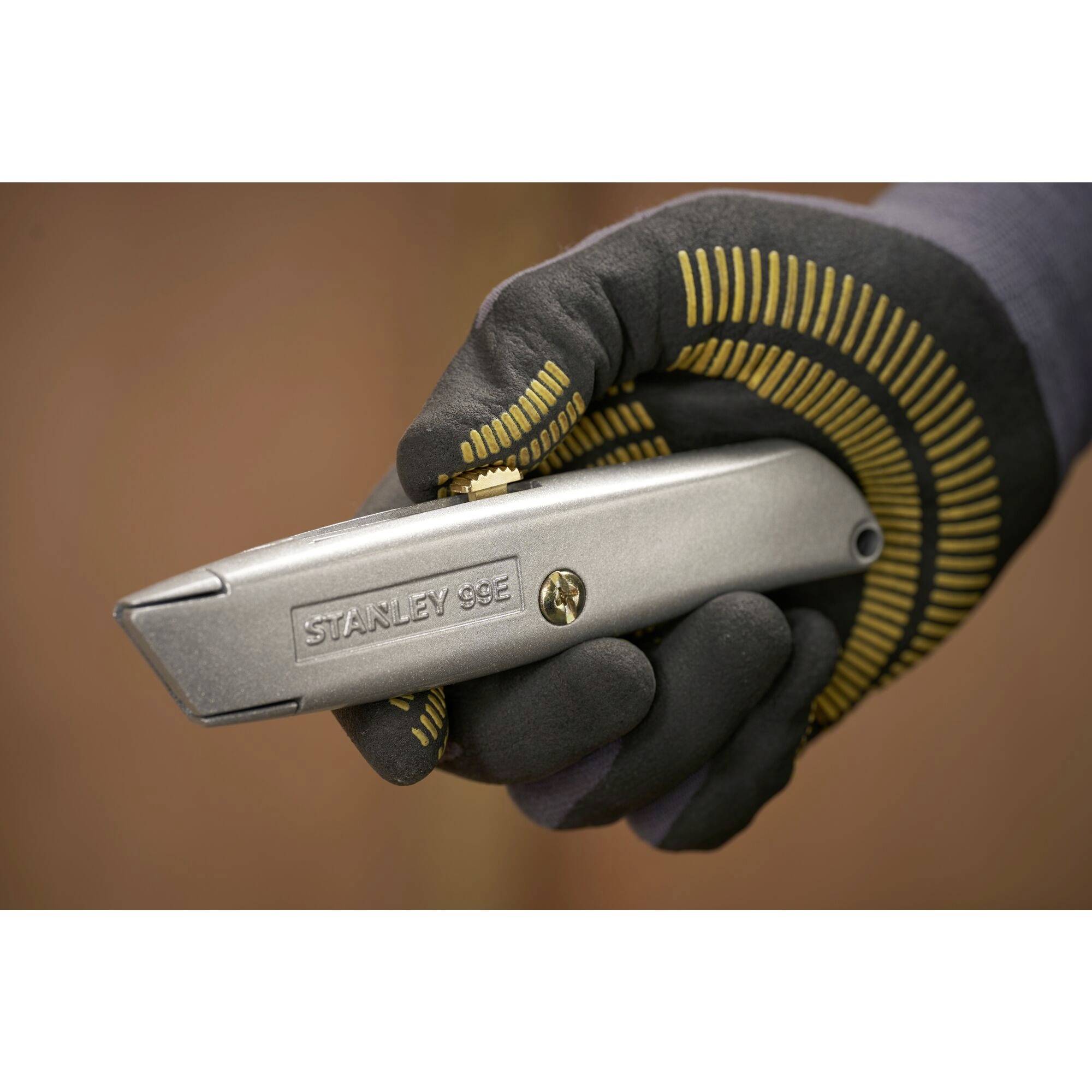 Eine bekleidete Hand hält ein Metall-Cutter-Messer. Das Messer zeigt die Aufschrift 'Stanley 99E', vorhanden ein Schraubmechanismus.