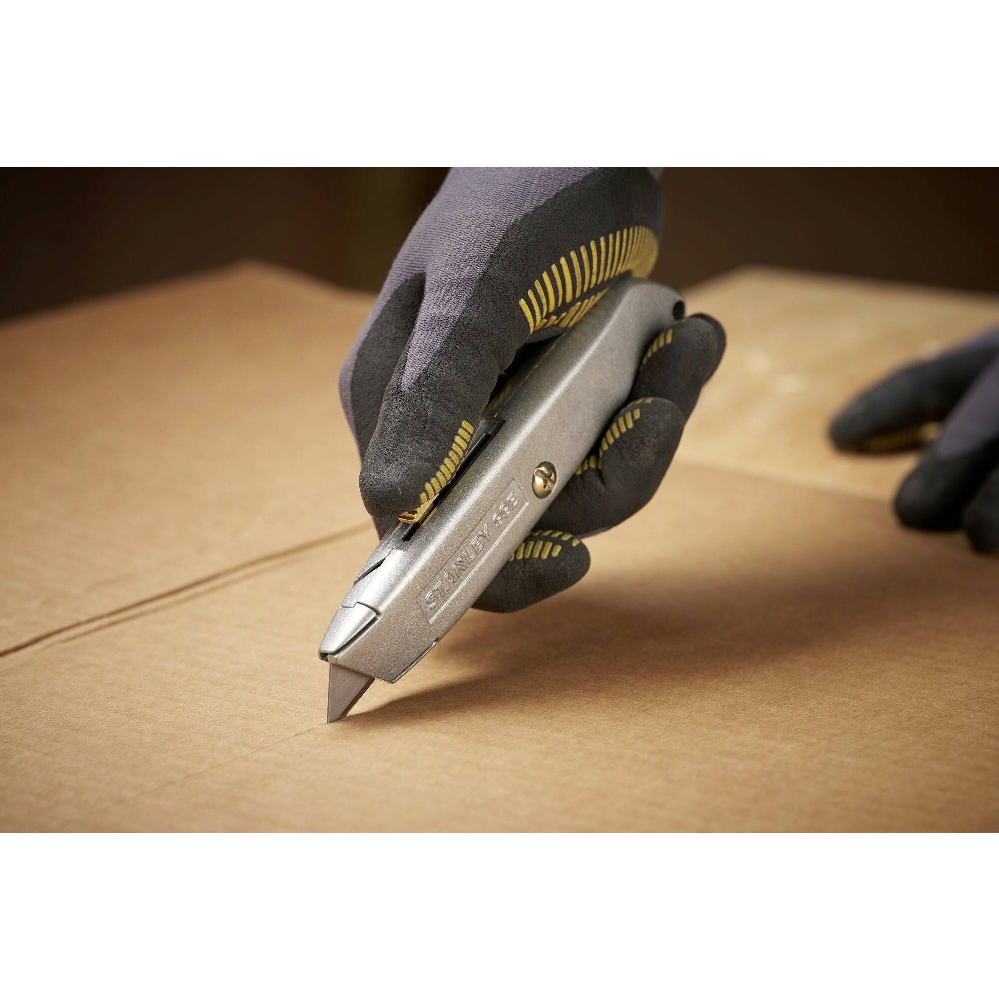 Eine Hand mit Handschuh hält ein silbernes Universalmesser und schneidet durch einen Pappkarton.