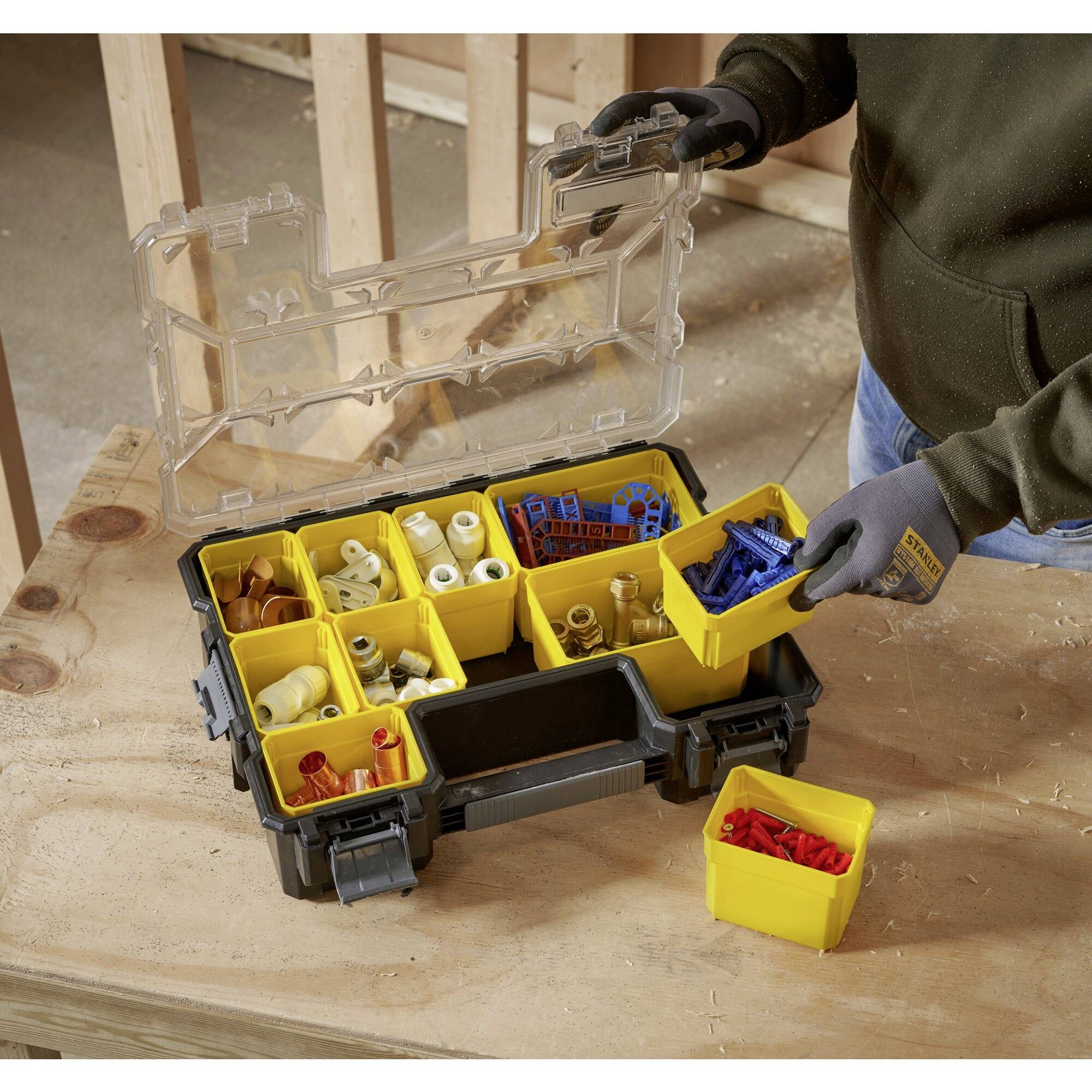 Eine geöffnete Werkzeugkiste mit gelben Fächern, gefüllt mit verschiedenen Befestigungselementen, liegt auf einem Holztisch. Eine Person nimmt einen Behälter heraus.