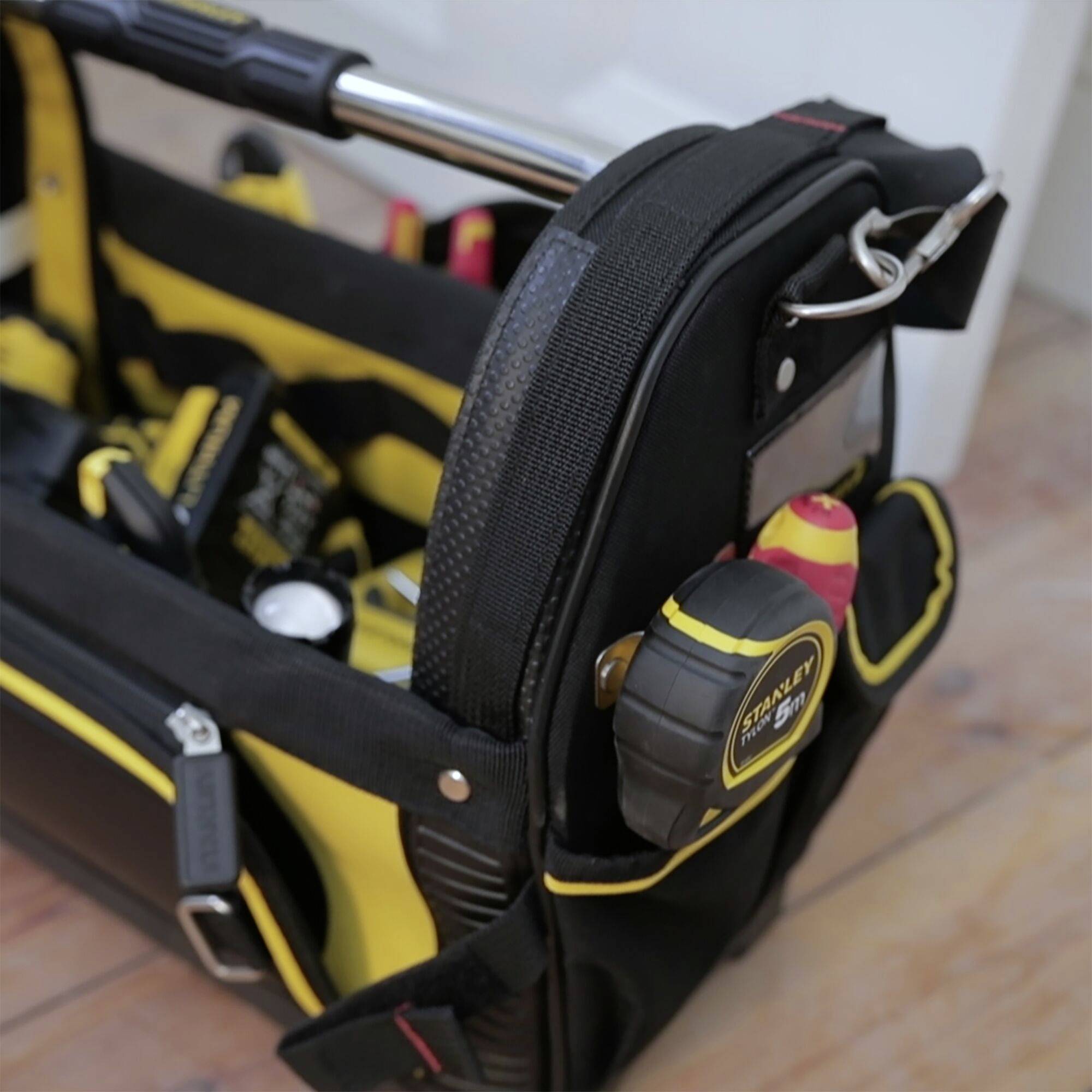 Werkzeugtasche mit gelbem und schwarzem Design auf Holzboden. Enthält verschiedenes Handwerkzeug wie Maßband, Schraubendreher und Zangen.