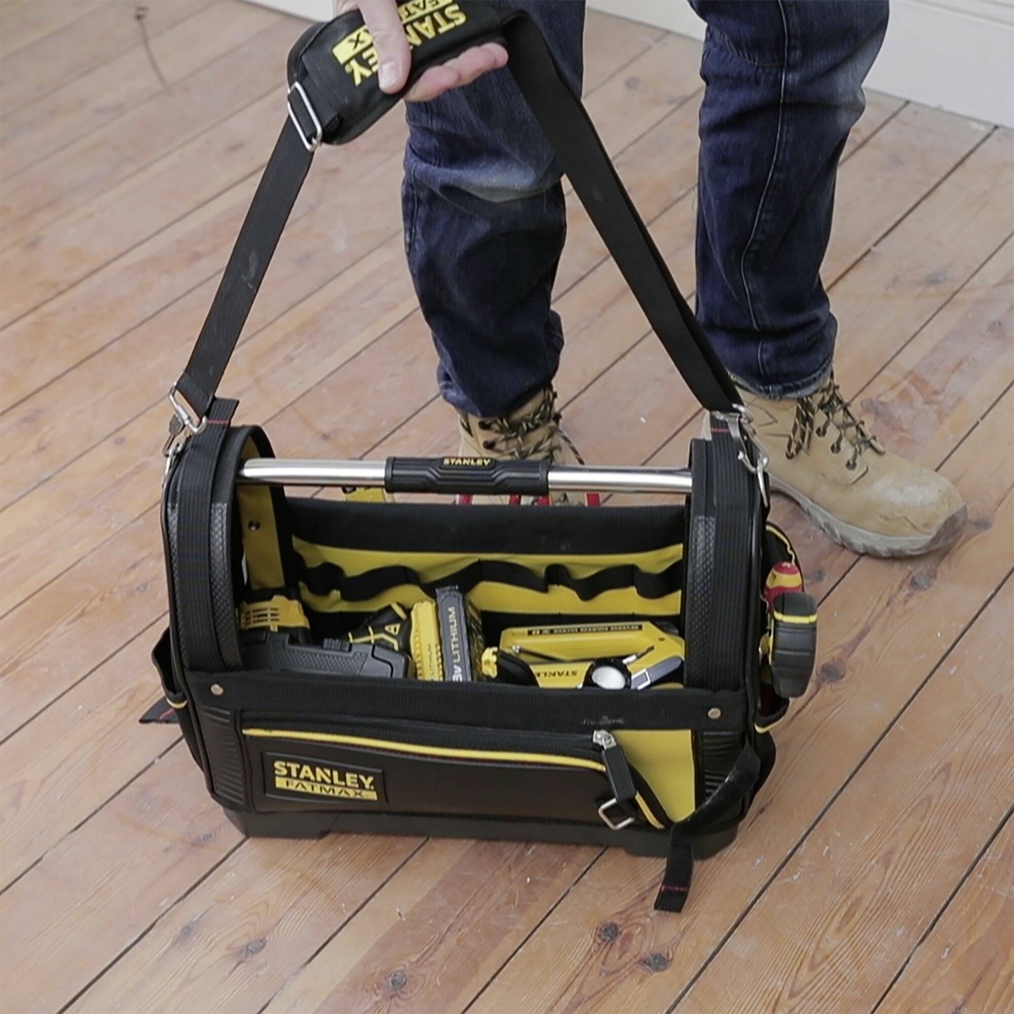 Eine Person trägt eine geöffnete Werkzeugtasche auf einem Holzboden. Die Tasche ist schwarz-gelb und enthält verschiedene Werkzeuge.