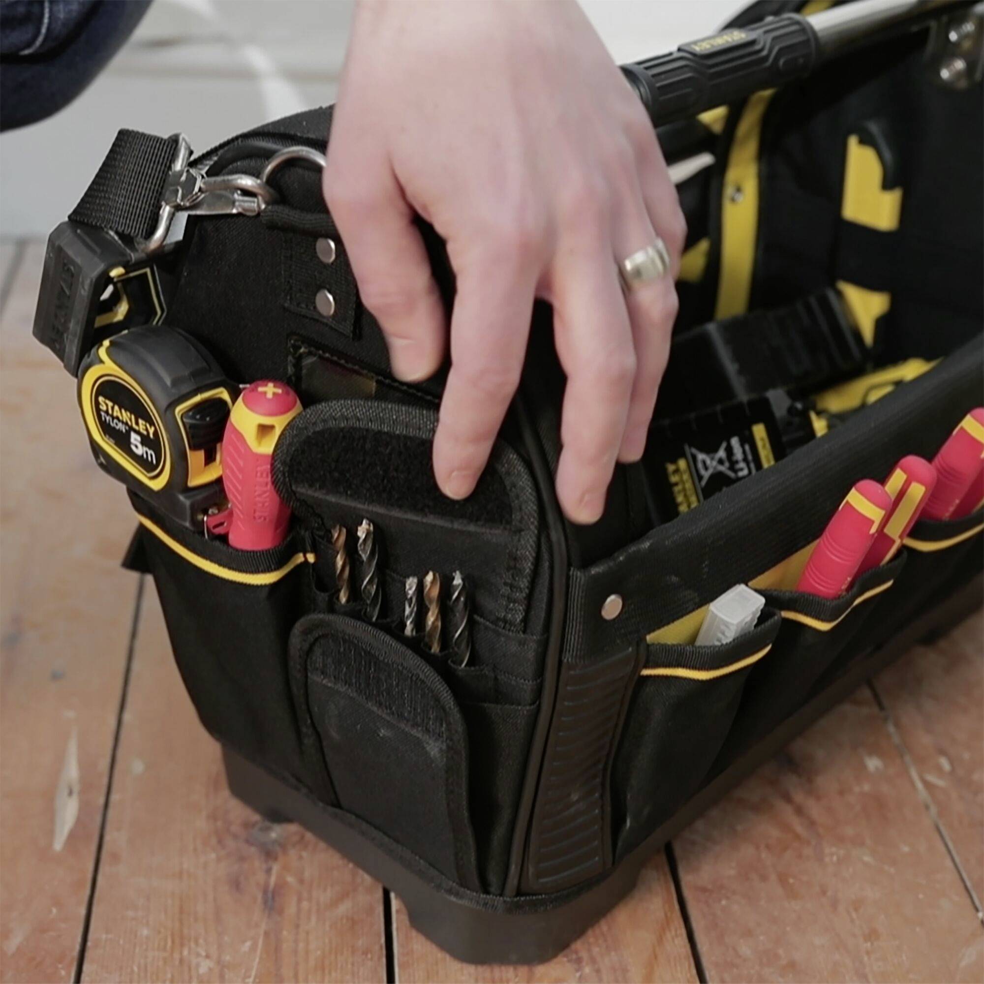 Eine Hand greift in eine offene Werkzeugtasche mit verschiedenen Schraubendrehern und Werkzeugen, auf einem Holzboden liegend.