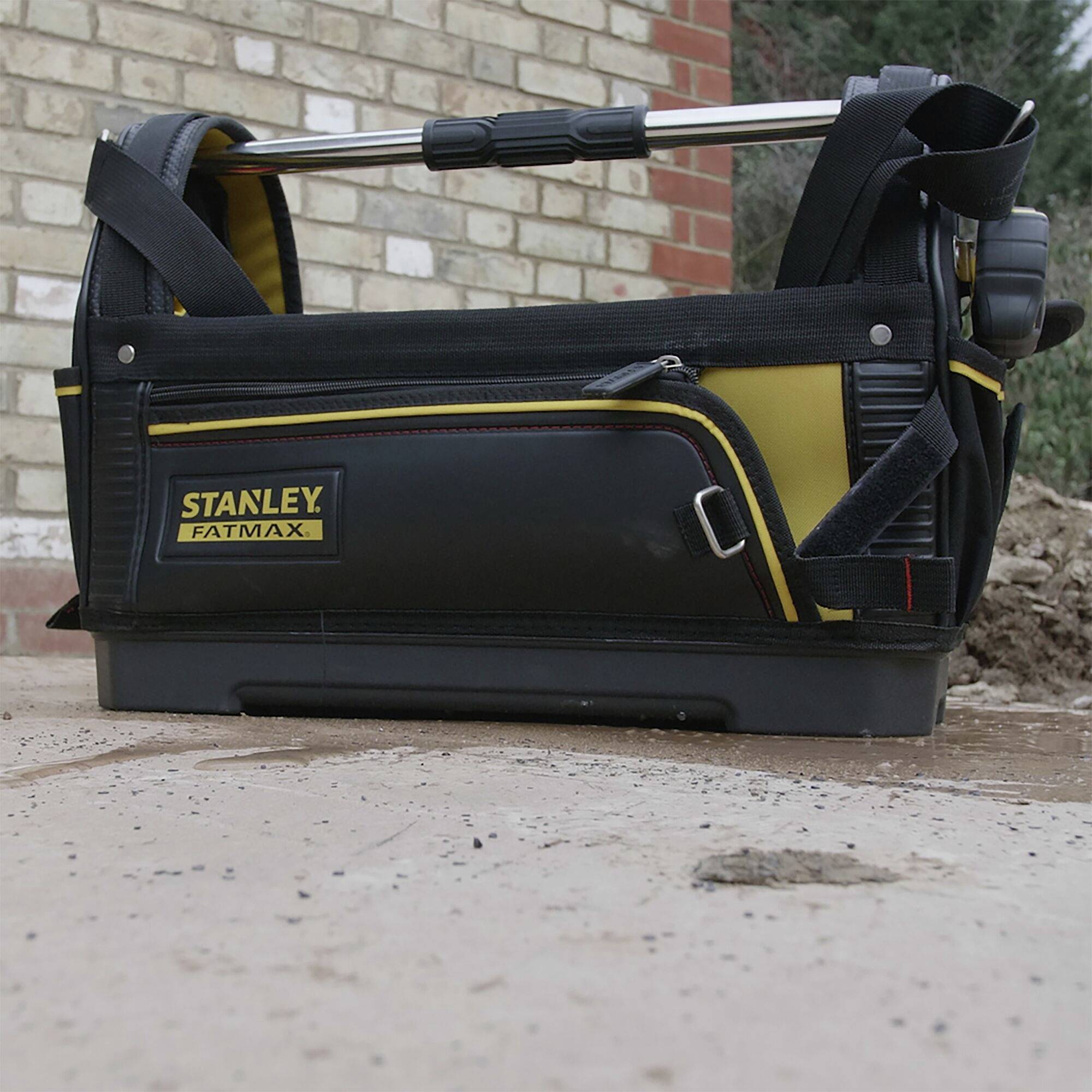 Eine schwarze und gelbe Werkzeugtasche von Stanley mit Trageriemen und Werkzeugen, auf einem gepflasterten Boden vor einer Ziegelmauer.