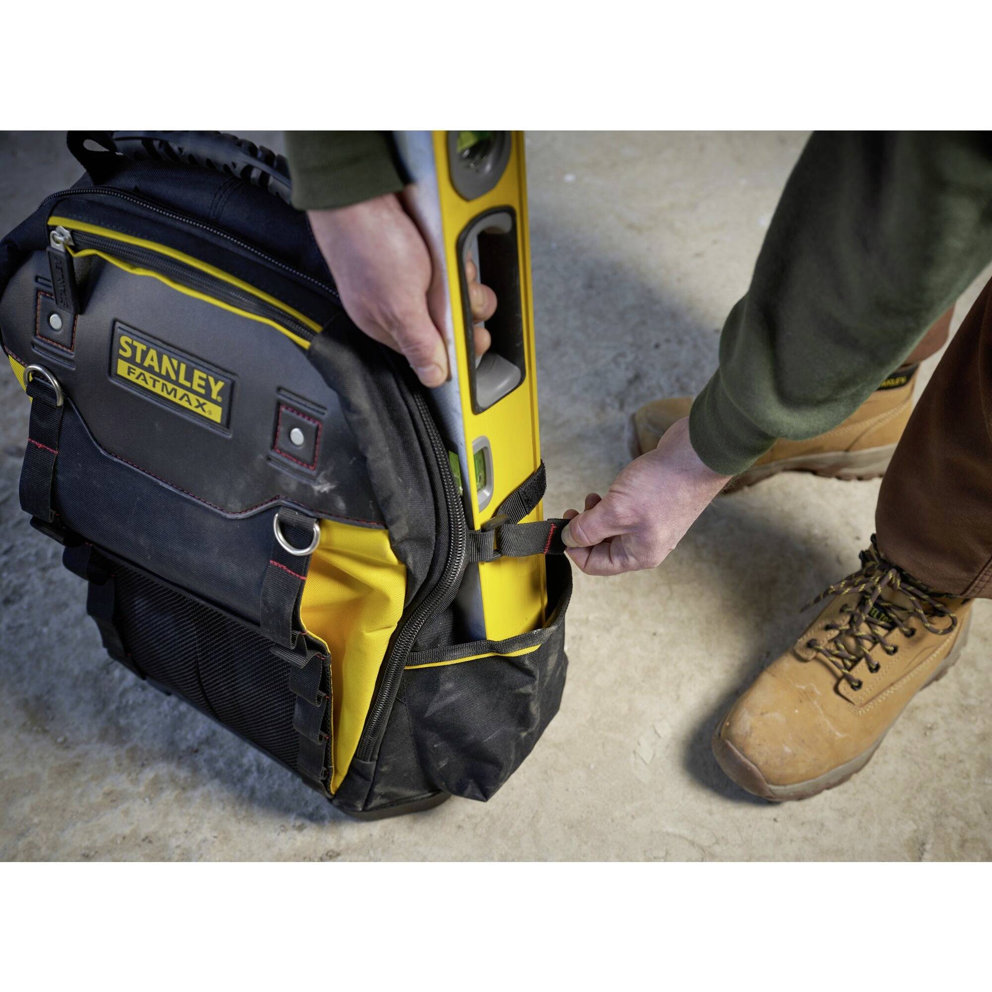 Eine Person hält eine Stanley-Tasche mit einem Wasserwaagen-Set. Die Tasche befindet sich auf einem grob strukturierten Boden, und die Person trägt robuste Stiefel.
