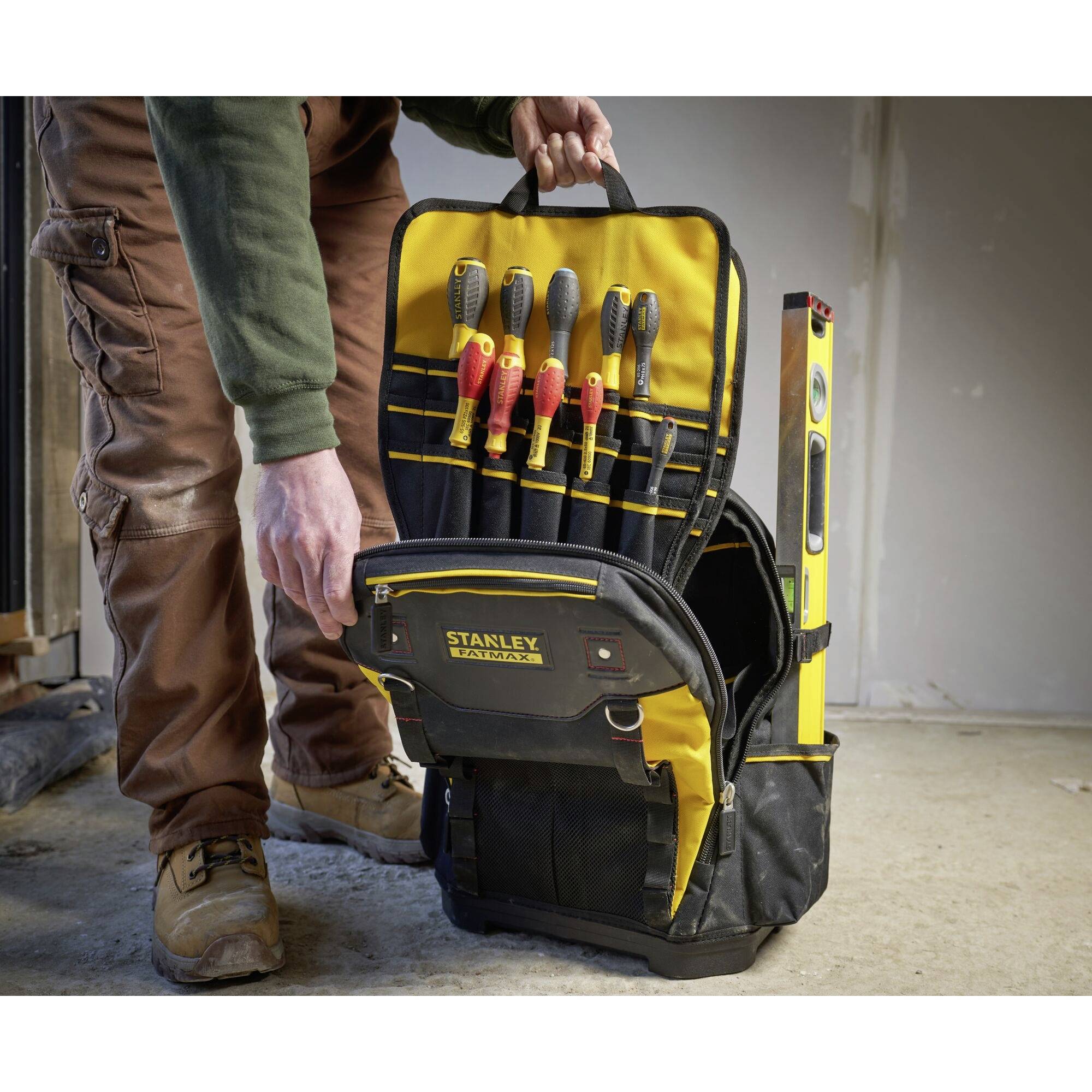 Handwerker steht mit einer schwarzen und gelben Werkzeugtasche, die Schraubenzieher und Wasserwaage enthält. Tasche offen und einsatzbereit.