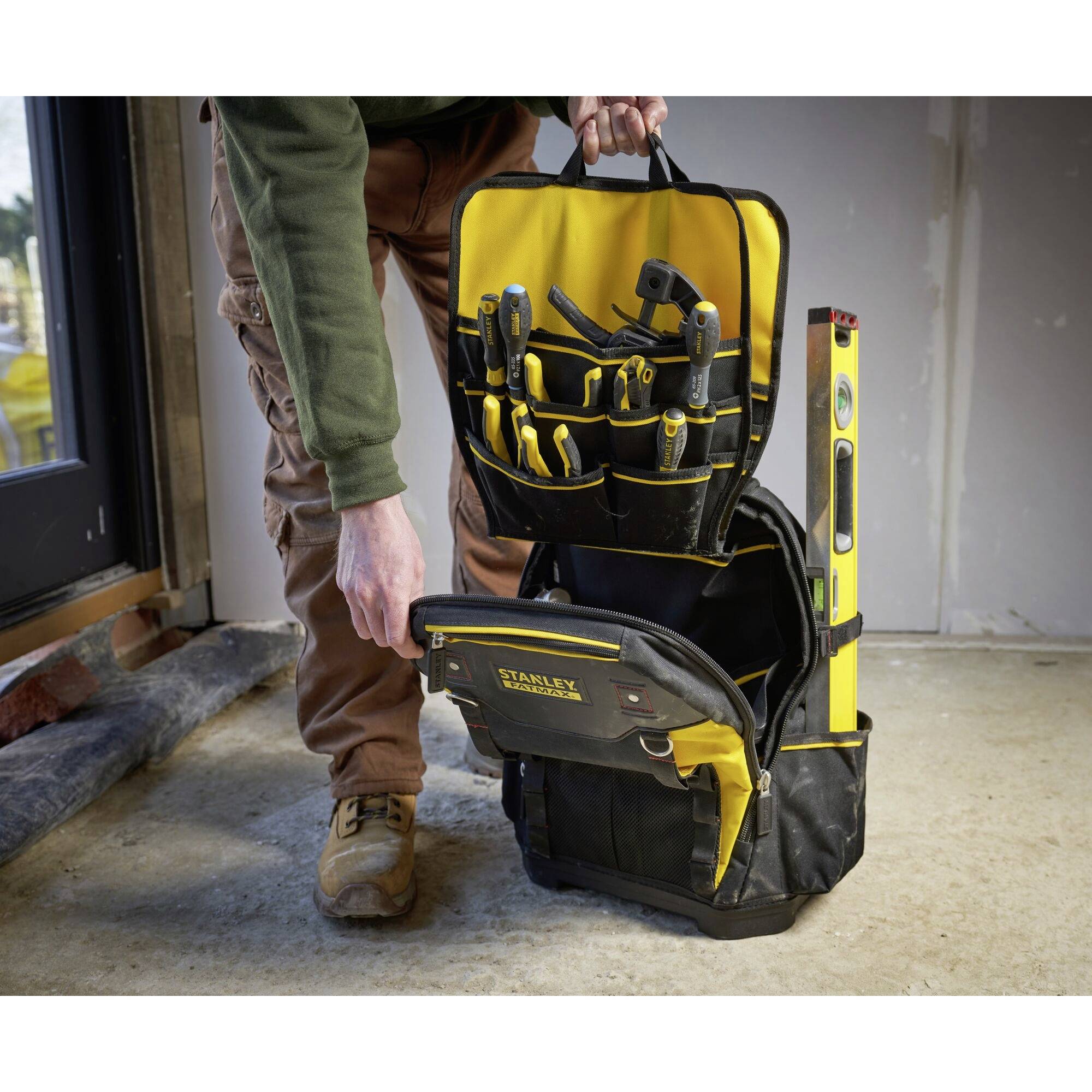 Eine Person hält eine Werkzeugtasche voller Werkzeuge. Eine Wasserwaage ragt aus der Tasche heraus. Die Szene findet in einem unfertigen Raum statt.