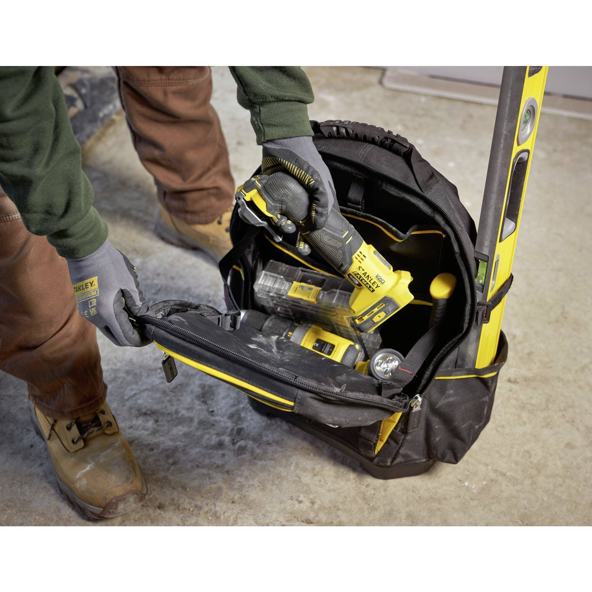 Eine Person packt Werkzeug in eine schwarze Tasche. Zu sehen sind Akku-Bohrschrauber und Wasserwaage auf einem Betonboden.