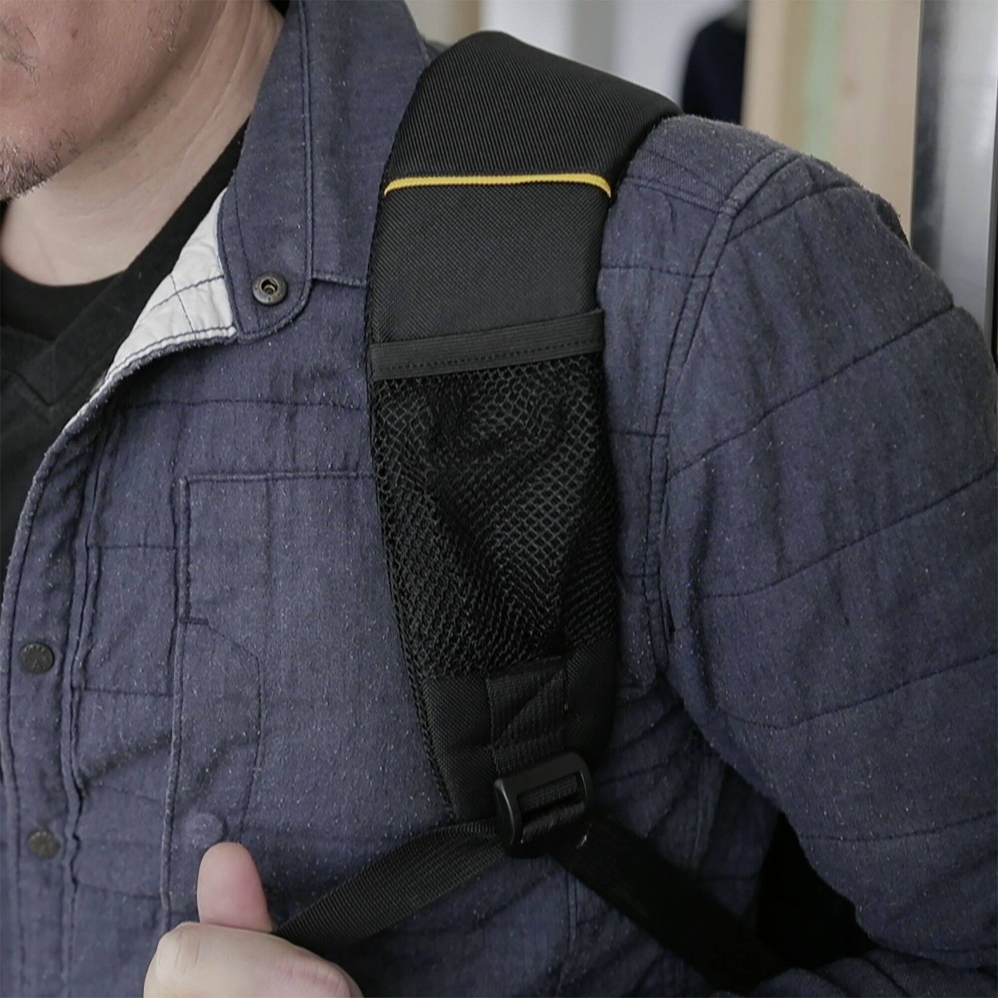 Person trägt eine dunkelblaue Jacke und einen schwarzen Rucksack. Der Riemen des Rucksacks ist deutlich sichtbar auf der Schulter.